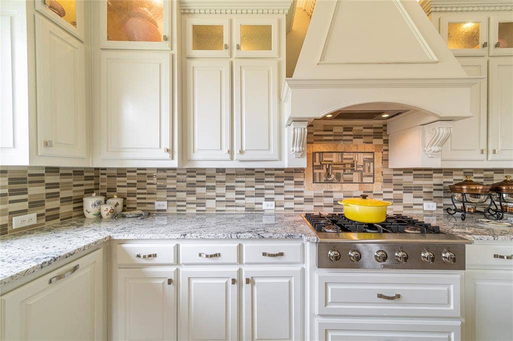13018 Seattle Slew Street, Frisco, TX 75035 White custom cabinetry with crown molding, stainless steel gas range, custom range hood, mosaic tile backsplash, and granite countertops