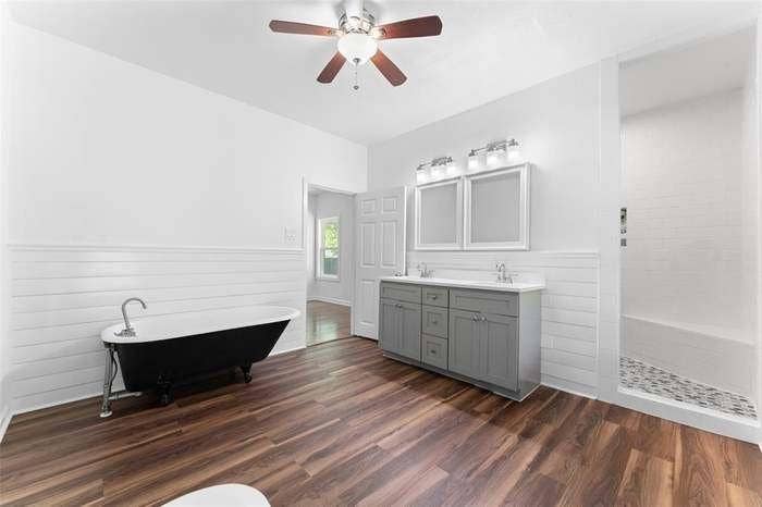 2405 Monroe Street, Commerce, TX 75428 Wood-finish flooring, dual vanity with gray cabinetry, white shiplap wall treatment, freestanding clawfoot tub, and a tiled shower with a built-in bench