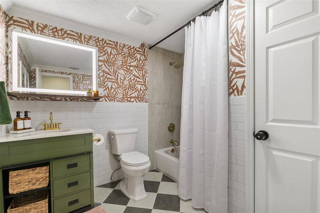 214 Short Street, McKinney, TX 75069 Bathroom featuring a green vanity with brass fixtures, an illuminated mirror, a toilet, a tiled shower, and patterned wallpaper
