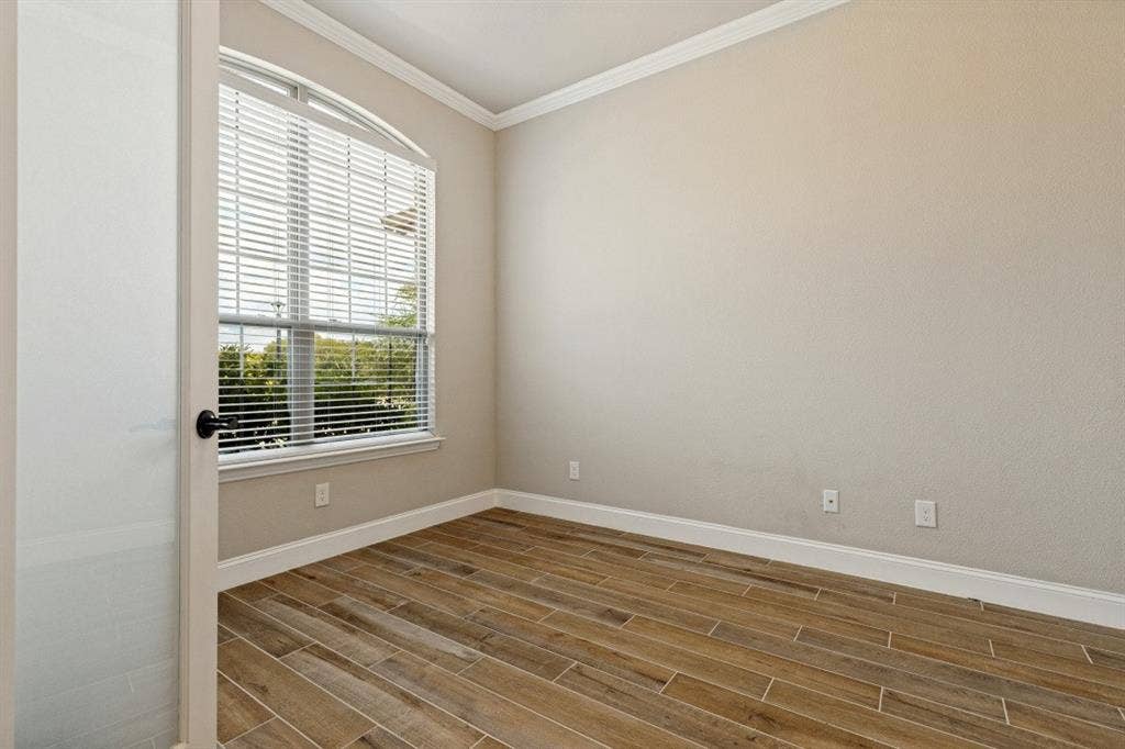 Arched window with blinds, wood-finish flooring, crown molding, white baseboards, and a solid door with a matte black handle