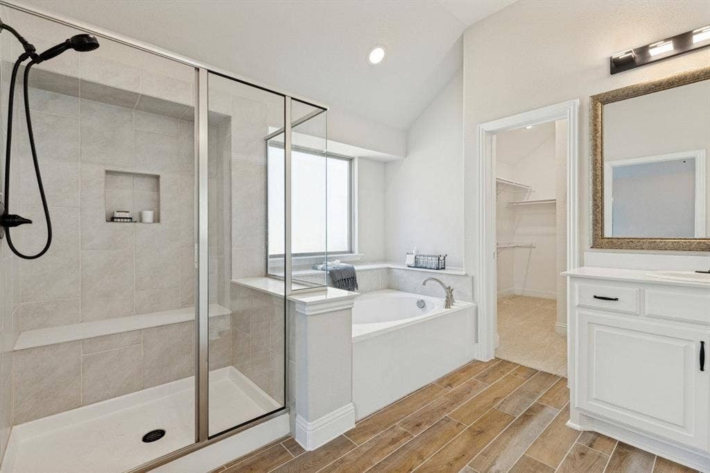 Walk-in shower with a built-in bench and recessed niche, soaking tub with window, wood-finish flooring, and a white vanity with a framed mirror