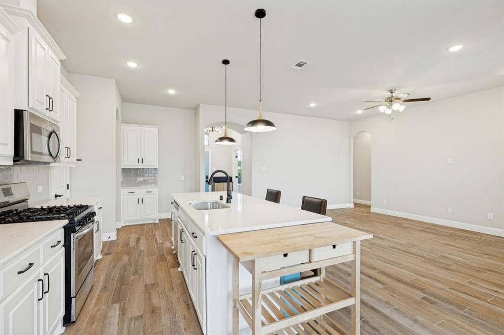 Kitchen featuring white cabinetry, stainless steel appliances, a center island with sink, pendant lighting, and wood-finish flooring also notice the wide-open living room. And recess lights