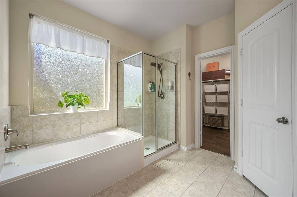 Soaking tub with tile surround, glass-enclosed shower, frosted window, and tile flooring