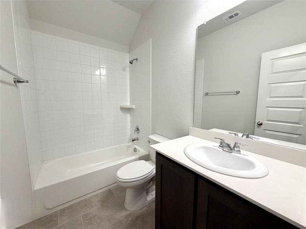 3806 Chicory Court, Melissa, TX 75454 This full guest bathroom showcases a clean and modern design, featuring a classic white bathtub complete with a stylish subway tile surround and a convenient built-in recessed shelf. The space is anchored by an elegant dark wood-finish vanity topped with a crisp white countertop, perfectly complemented by high-quality tiled flooring that adds both durability and visual appeal to the room.
