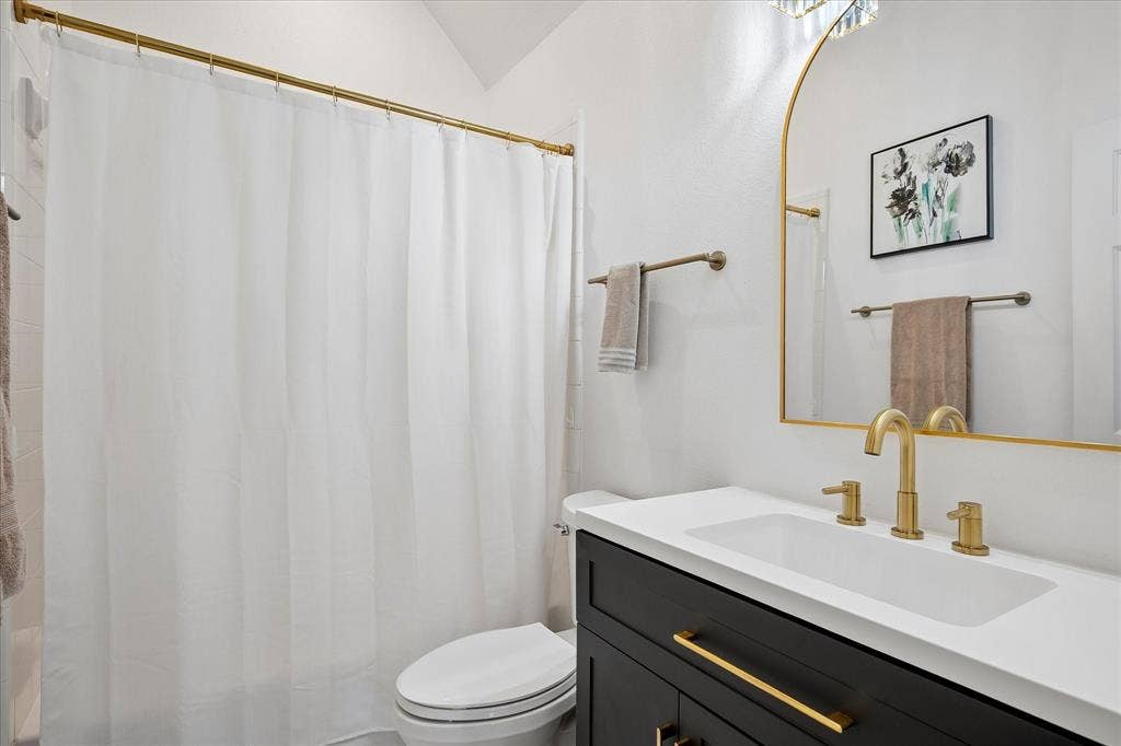 Bathroom featuring a dark wood-finish vanity with gold-tone hardware, white countertop with integrated sink, gold-tone faucet, and an arched gold-tone framed mirror