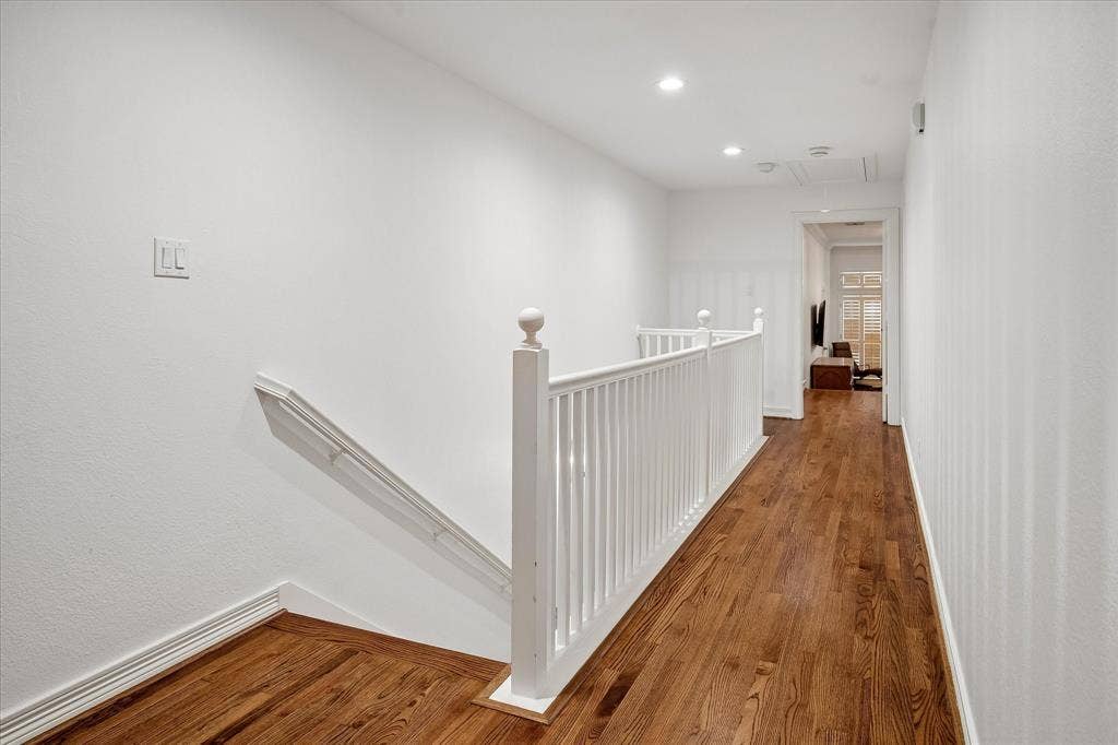 Hardwood flooring throughout the hallway, white painted walls, white spindle railing, and recessed ceiling lighting