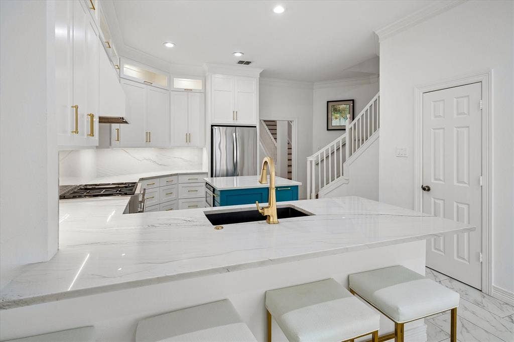 White cabinetry with gold-finish hardware, a large kitchen island with a gold-finish faucet, a contrasting blue island base, a built-in stainless steel refrigerator, and a recessed cooktop with a matching range hood