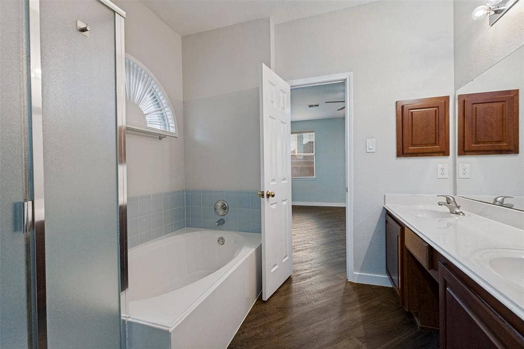 4215 Maidstone Drive, Garland, TX 75043 Primary bathroom featuring a built-in tub, a frosted glass shower enclosure, a double vanity with an integrated countertop, and wood-finish flooring