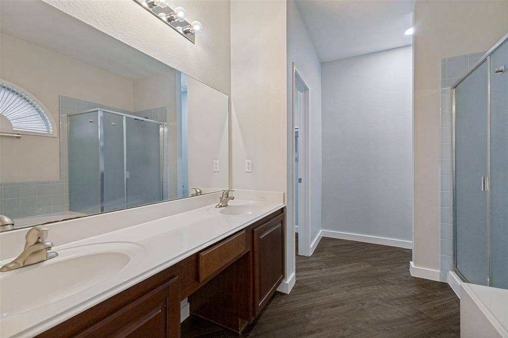 4215 Maidstone Drive, Garland, TX 75043 Primary bathroom featuring a built-in tub, a frosted glass shower enclosure, a double vanity with an integrated countertop, and wood-finish flooring expansive mirror, and a three-light fixture