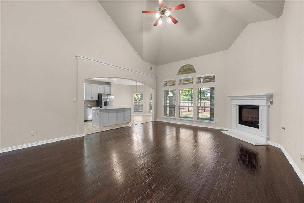 Vaulted ceiling with a ceiling fan, dark wood-finish flooring, white fireplace with hearth, arched entryway, and multiple windows