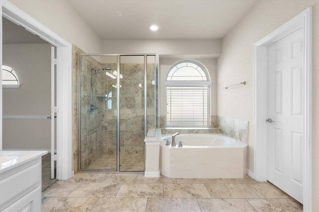 Tiled flooring throughout, corner soaking tub with deck-mounted fixtures, separate glass-enclosed shower with tiled walls, and an arched transom window with blinds