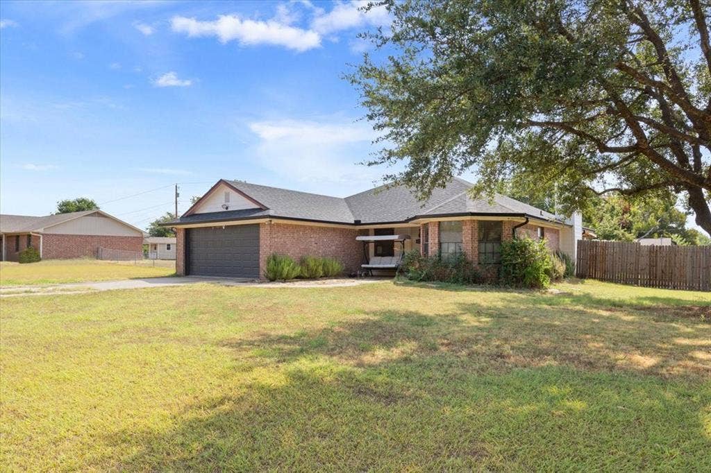 Brick exterior with a dark shingle roof, attached garage, mature front yard tree, and a wood privacy fence