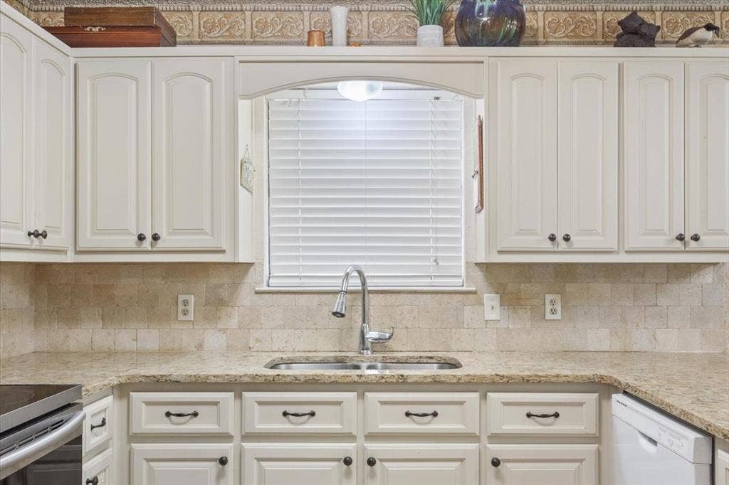 White paneled cabinetry, stone countertops, stainless steel double basin sink with a gooseneck faucet, and a stone tile backsplash