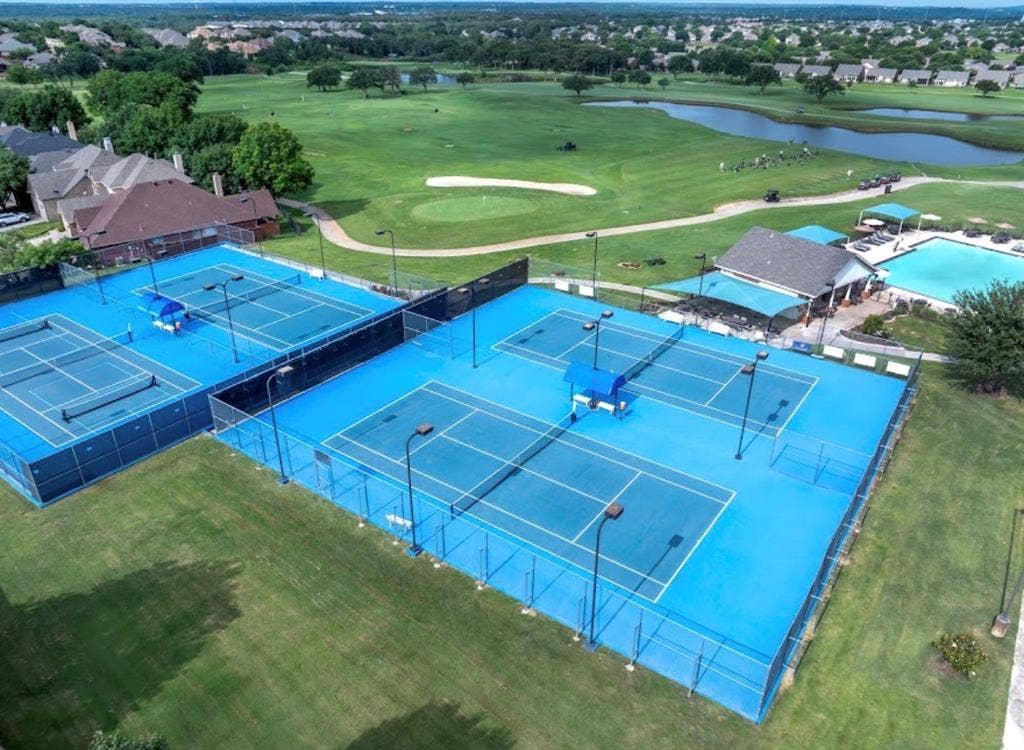 Multiple tennis courts with blue surfacing, surrounded by green landscaping