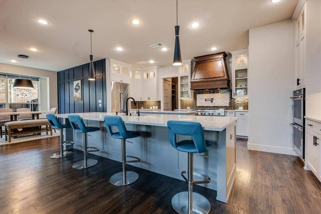 4102 Parkview Boulevard, Celina, TX 75009 Kitchen featuring an expansive island with a light-toned countertop, wood-finish flooring, white cabinetry, a built-in double oven, and a decorative range hood