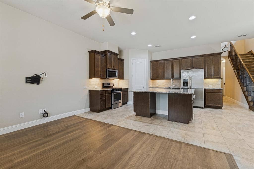 Open-concept layout featuring wood-flooring, a kitchen island with granite countertop, stainless steel appliances, and dark wood cabinetry