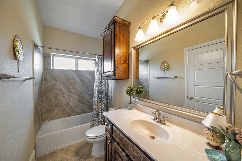13018 Seattle Slew Street, Frisco, TX 75035 Downstairs 2nd bath Vanity with an integrated sink and brushed nickel faucet, large framed mirror with overhead lighting, a built-in wood-finish cabinet, tiled shower-tub combination, and a window above the tub
