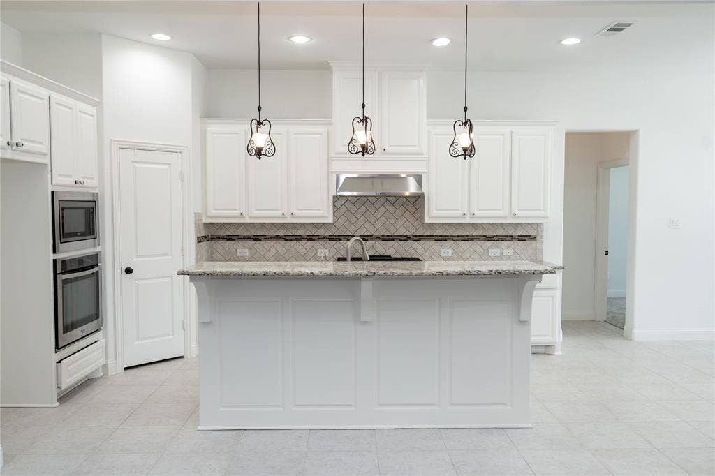 Kitchen featuring a central island with an integrated sink, white cabinetry, a herringbone tile backsplash, stainless steel appliances, and pendant lighting