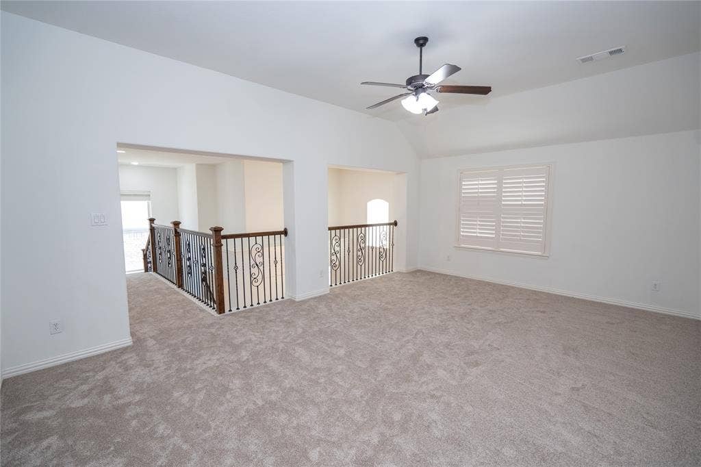Carpeted loft space featuring a vaulted ceiling, ceiling fan with light, window with plantation shutters, and ornate metal railings