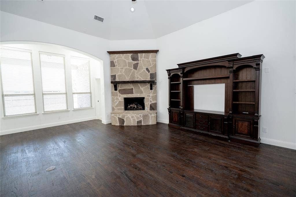 Vaulted ceiling, stone-clad fireplace with mantel, dark wood-finish flooring, built-in entertainment center, and multiple windows with blinds