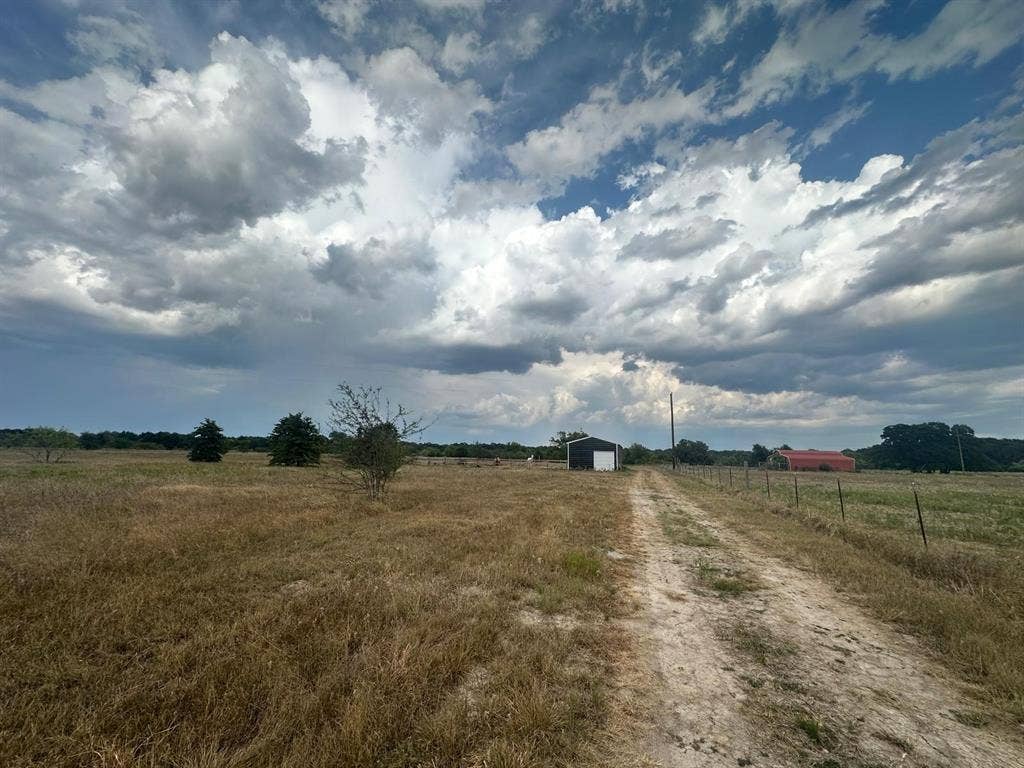 12496 County Road 2148, Kemp, TX 75143 Expansive open field with a dirt pathway, featuring a black and white barn, a red barn, and mature trees