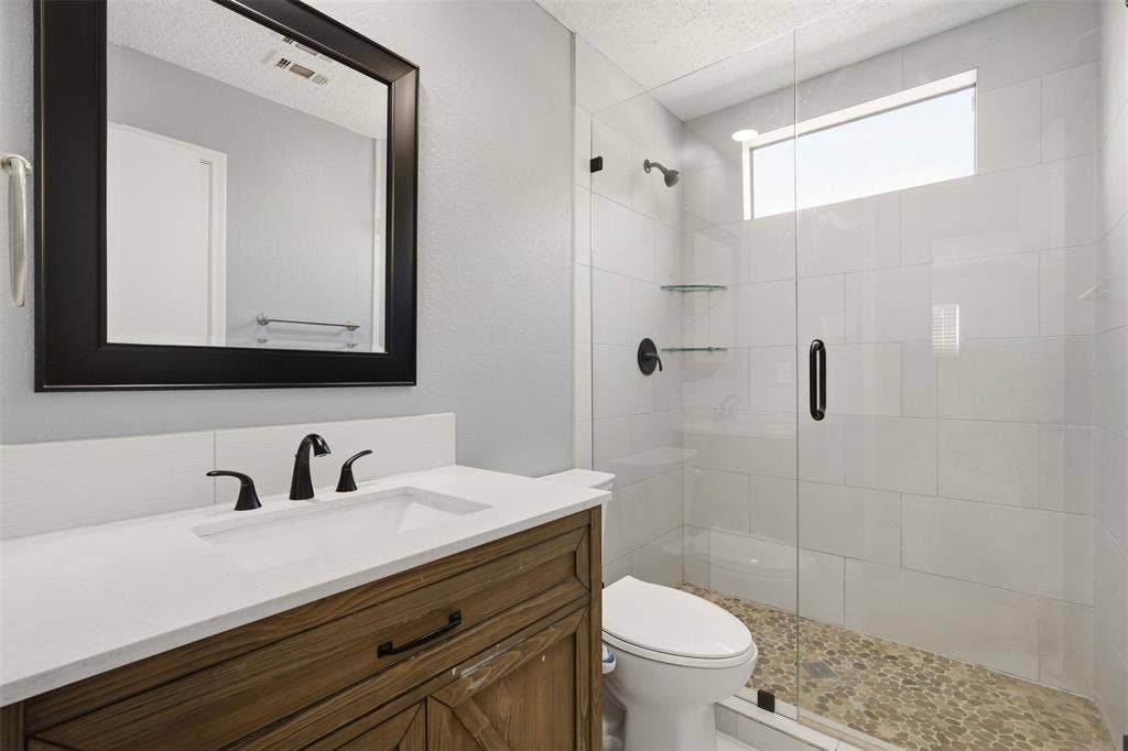 1512 Woodcreek Drive, Richardson, TX 75082 Wood-finish vanity with an integrated sink and matte black faucet, framed mirror, and a glass-enclosed shower with pebble flooring and a tiled surround