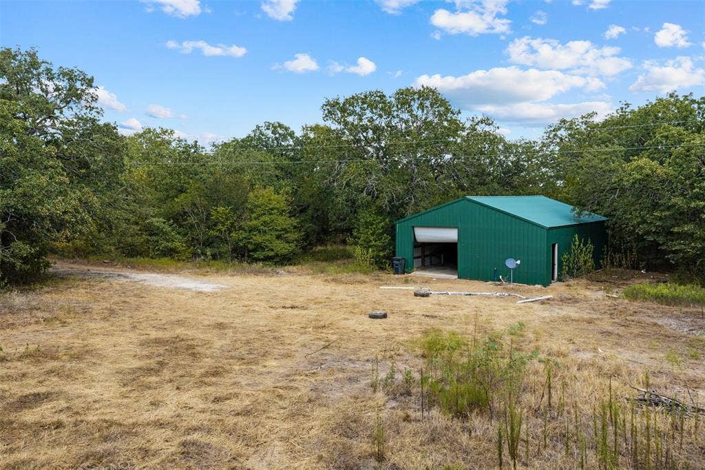 172 M Burton Street, Riesel, TX 76682 Green metal outbuilding with a roll-up door and a side entry, surrounded by mature trees
