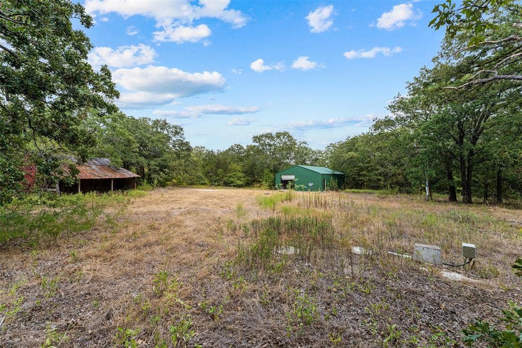 172 M Burton Street, Riesel, TX 76682 Expansive open field with scattered trees, featuring a red structure with a covered porch and a green metal building