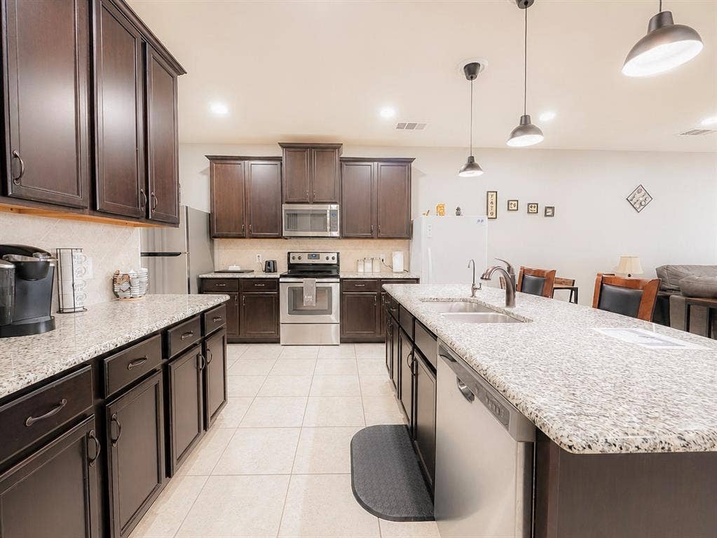 Kitchen featuring dark wood-finish cabinetry, light speckled countertops, stainless steel appliances, a double basin sink, and pendant lighting