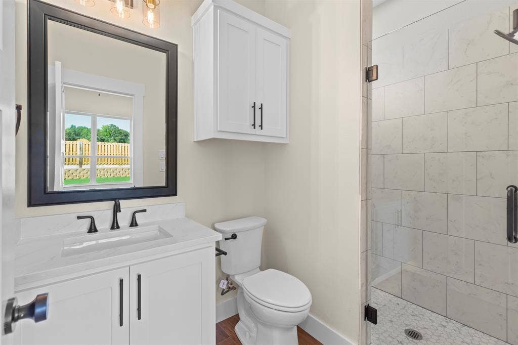 White vanity with an integrated sink and black faucet, framed mirror, built-in storage cabinet, white toilet, and a glass-enclosed shower with tiled walls and a mosaic floor