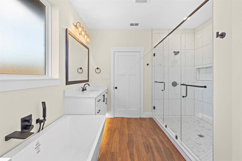 Spacious bathroom featuring a freestanding soaking tub with wall-mounted fixtures, a glass-enclosed shower with tile surround, a single vanity with a white countertop, and wood-finish flooring