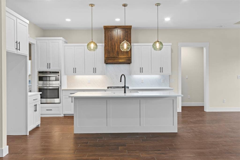 Kitchen featuring white shaker cabinetry, stainless steel double wall ovens, a center island with sink, wood-finish flooring, and pendant lighting