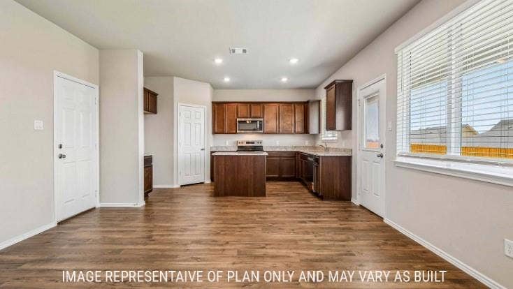 Open-concept kitchen and dining area featuring wood-finish flooring, wood cabinetry with stainless steel appliances, a centered island with seating, recessed lighting, and abundant natural light from large windows overlooking the landscape