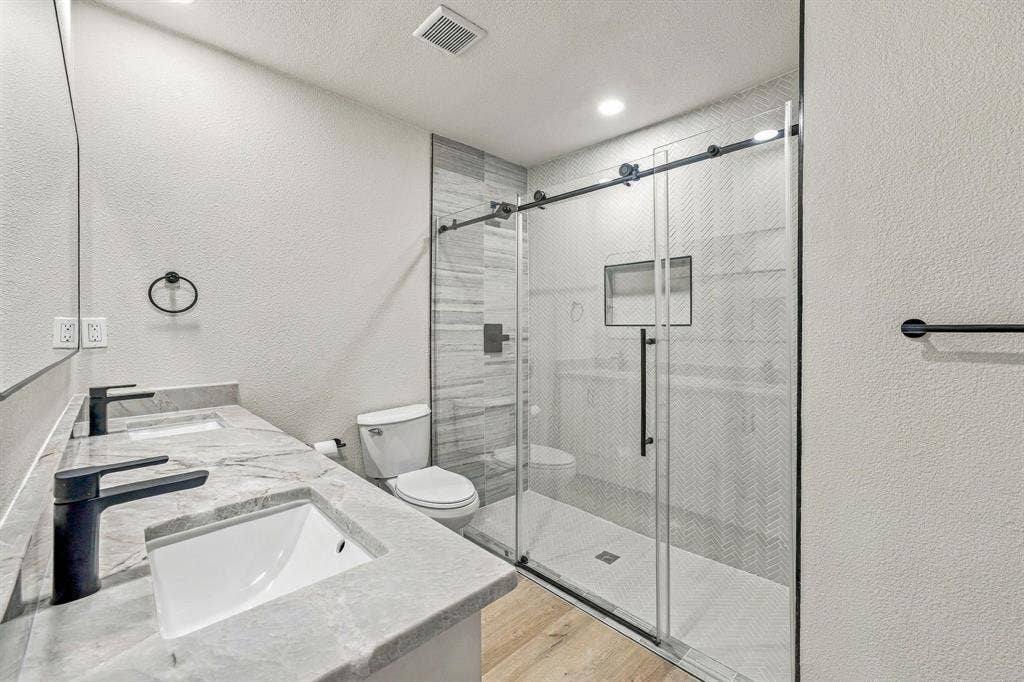 13706 Janwood Lane, Farmers Branch, TX 75234 Double vanity featuring rectangular undermount sinks, stone-finish countertops, matte black fixtures, and a walk-in shower with sliding glass door and herringbone-pattern tile surround
