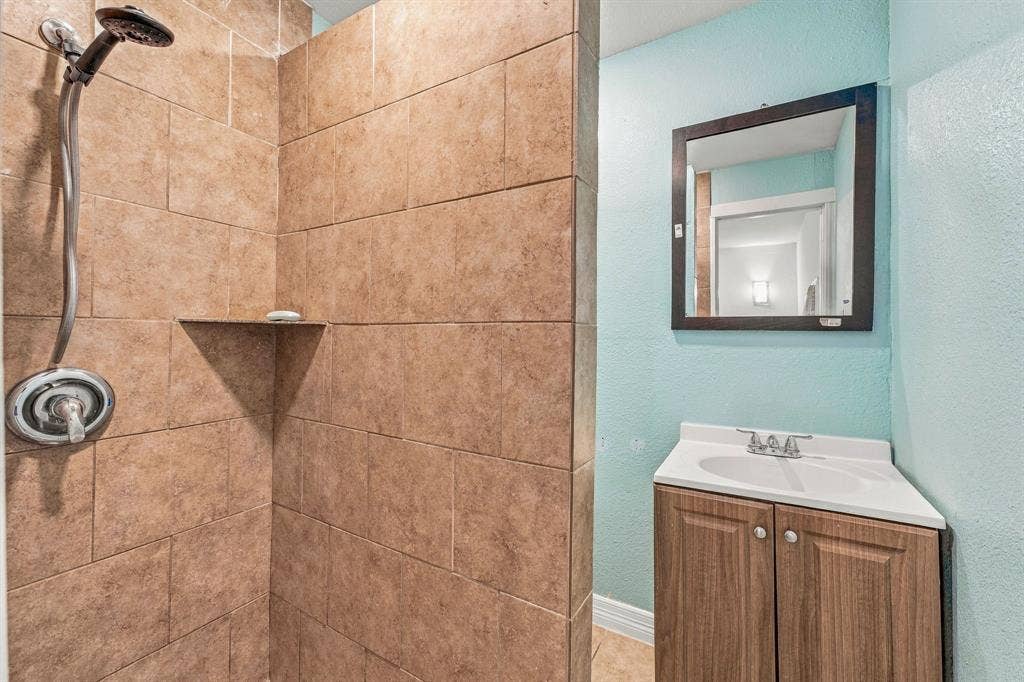 13706 Janwood Lane, Farmers Branch, TX 75234 Walk-in shower with floor-to-ceiling tile surround and built-in stone corner shelf, featuring a chrome adjustable showerhead, wood-finish vanity cabinet, integrated sink, and framed wall mirror