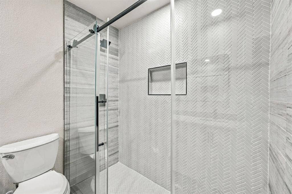 13706 Janwood Lane, Farmers Branch, TX 75234 Walk-in shower featuring herringbone-patterned wall tile, glass sliding door with matte black hardware, built-in recessed niche, and horizontal-set tile accent wall