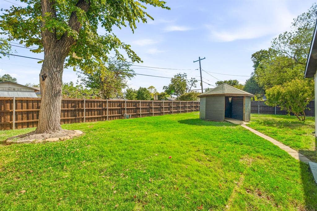 13706 Janwood Lane, Farmers Branch, TX 75234 Backyard featuring a manicured lawn, perimeter wood fencing, mature shade tree with stone edging, and a detached hexagonal structure with a paved walkway
