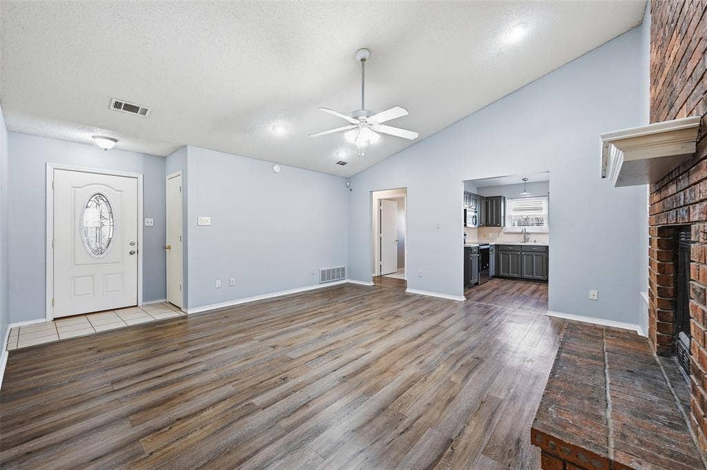 6421 Fairview Drive, Watauga, TX 76148 Vaulted ceiling with a ceiling fan, wood-finish flooring, brick fireplace with a mantel, and a kitchen with dark cabinetry