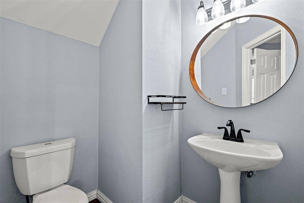 14592 Aspen Court, Addison, TX 75001 Pedestal sink, matte black faucet, wall-mounted oval mirror, three-light vanity fixture, and a toilet