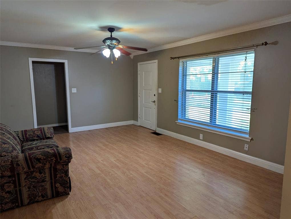 111 Murchison Street, Farmersville, TX 75442 Spacious living room featuring wood-look flooring, neutral wall colors, crown molding, and a ceiling fan with lighting. Large front window welcomes natural light, creating a comfortable space ready for your personal touches!