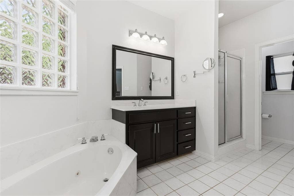 1926 Vintage Drive, Corinth, TX 76210 White tile flooring, corner jetted tub, glass block window, dark wood-finish vanity with countertop, and glass-enclosed shower
