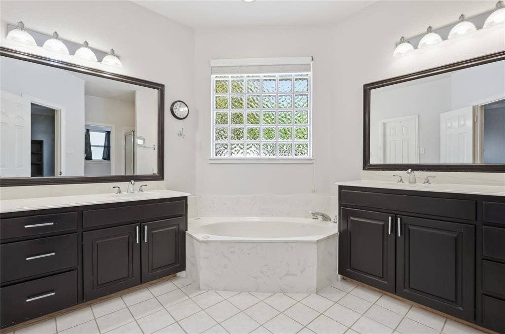 1926 Vintage Drive, Corinth, TX 76210 White tile flooring, dual dark wood-finish vanities with light countertops, large framed mirrors, and a built-in soaking tub with a tiled surround