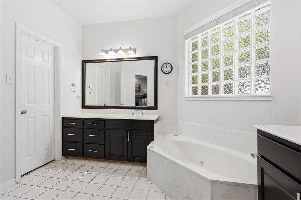 1926 Vintage Drive, Corinth, TX 76210 Expansive bathroom featuring a corner soaking tub, a wide vanity with a single basin, dark wood-finish cabinetry, a large framed mirror, and a privacy glass block window