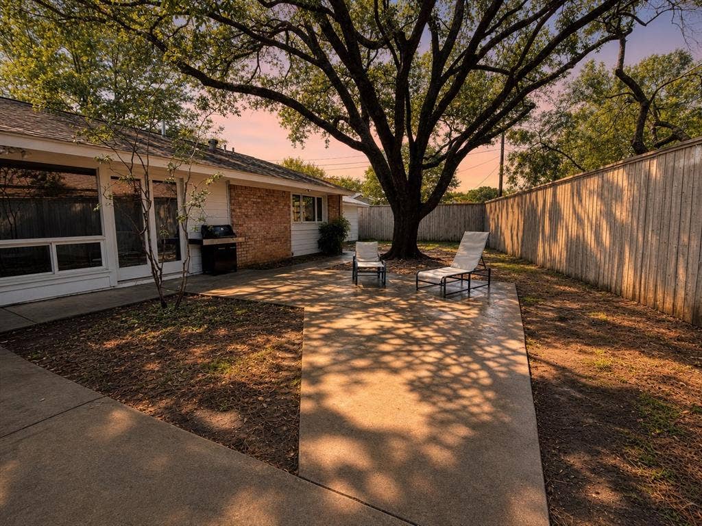 3401 Hanover Lane, Irving, TX 75062 Private backyard featuring an expansive concrete patio, mature trees providing natural shade, a wood privacy fence, and brick exterior accents on the home