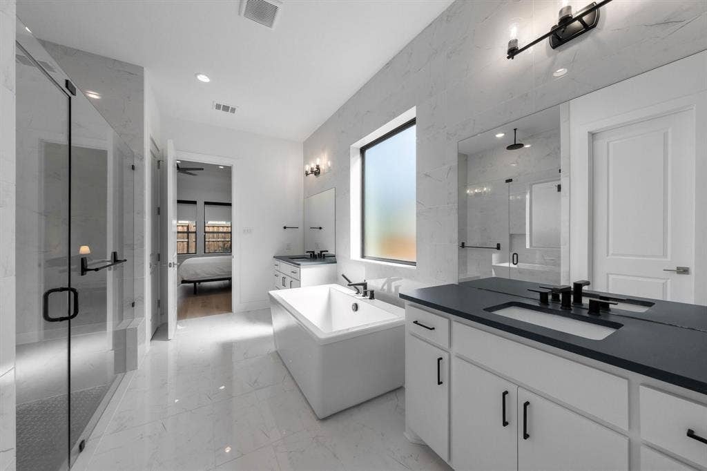 Spacious bathroom featuring white tiled flooring, a large freestanding soaking tub, a glass-enclosed shower, a double vanity with dark countertops, and a rectangular window
