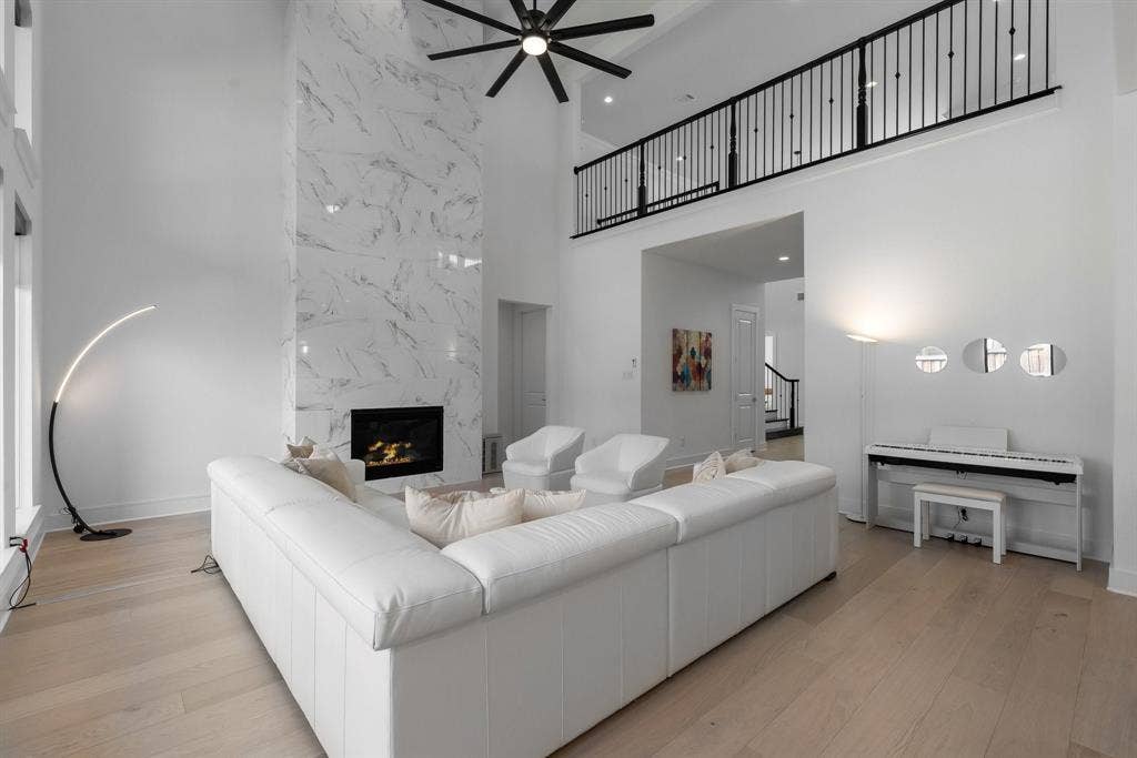 Vaulted ceiling with black ceiling fan, two-story fireplace with white marble-patterned surround, light wood-finish flooring, and black metal open-rail balcony
