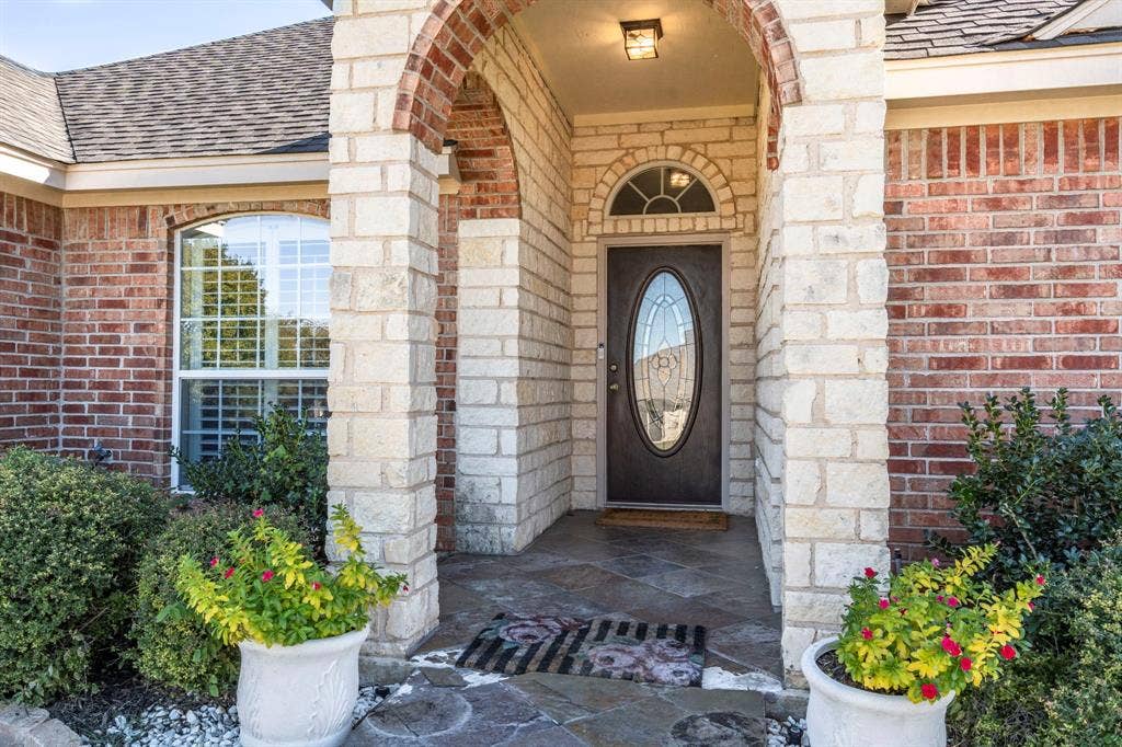 11701 Cambria Court, Fort Worth, TX 76008 Arched entryway featuring brick and stone masonry, a dark paneled door with an oval glass insert, a transom window, and a stone-tiled porch