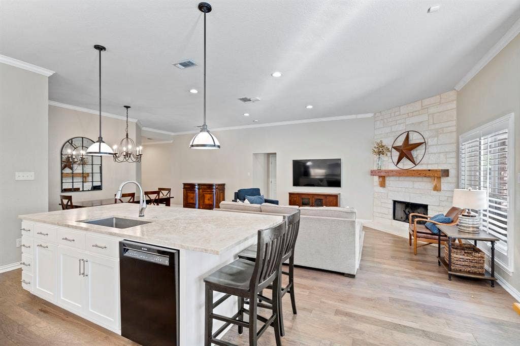 11701 Cambria Court, Fort Worth, TX 76008 Spacious kitchen island with a built-in sink and dishwasher, pendant lighting, stone-clad fireplace with a wood mantel, and wood-finish flooring