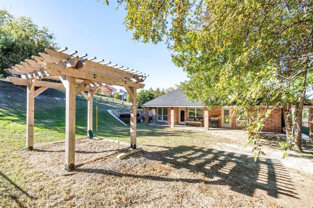 11701 Cambria Court, Fort Worth, TX 76008 Wood pergola over a gravel base, expansive lawn, mature foliage, brick exterior, and covered patio