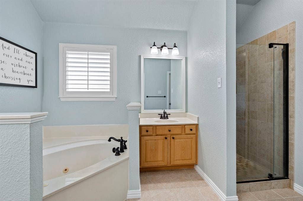 11701 Cambria Court, Fort Worth, TX 76008 Corner soaking tub, wood vanity with a light-colored countertop, framed mirror, three-light vanity fixture, and a tiled walk-in shower with a glass enclosure